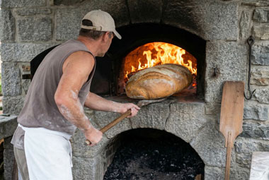 Ein Bäcker holt mit einem langen Holzschieber ein riesiges Brot aus einem Steinofen