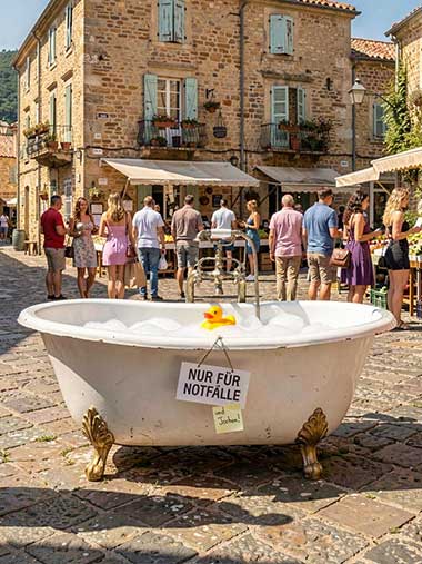 Volle Badewanne mit Quietscheentchen in der Mitte eines geschäftigten Marktplatzes. Zwei MÄnner in Bademänteln stehen davor. Auf dem Schild an der Badewanne steht 'Nur für Notfälle - und Jochen!'